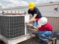 Two workers on the roof of a building working on the air conditioning unit.